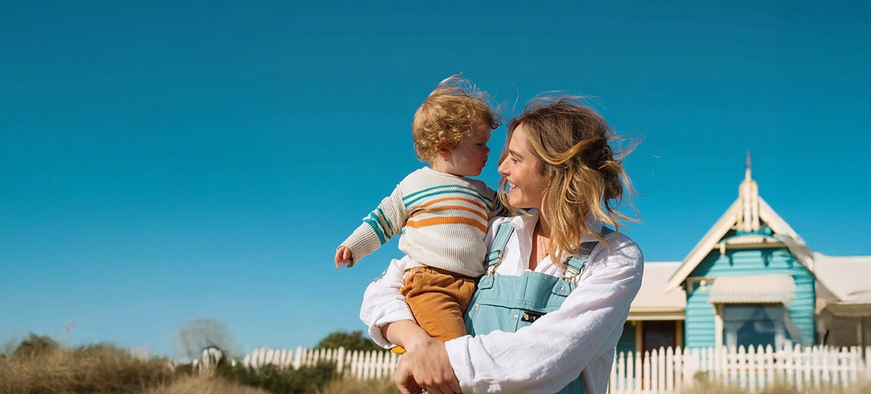Smiling woman holding a toddler with curly hair outdoors near a white picket fence and a blue house under a clear blue sky.