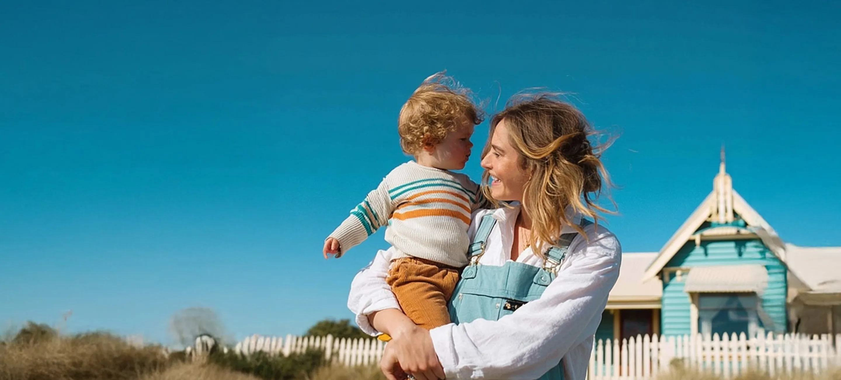 Smiling woman holding a toddler with curly hair outdoors near a white picket fence and a blue house under a clear blue sky.