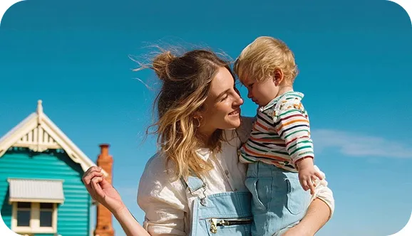 Smiling woman in denim overalls holding a toddler in striped shirt under clear blue sky with house in background.