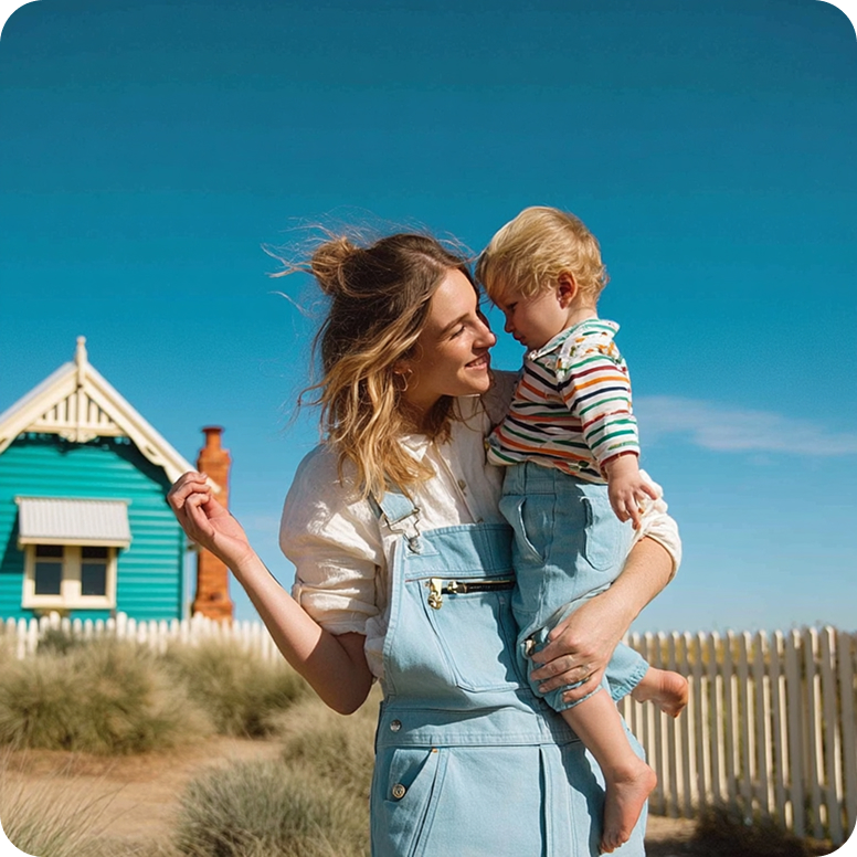 Smiling woman in light blue overalls holding a toddler in striped shirt and denim shorts outdoors with a blue house and white fence in the background.