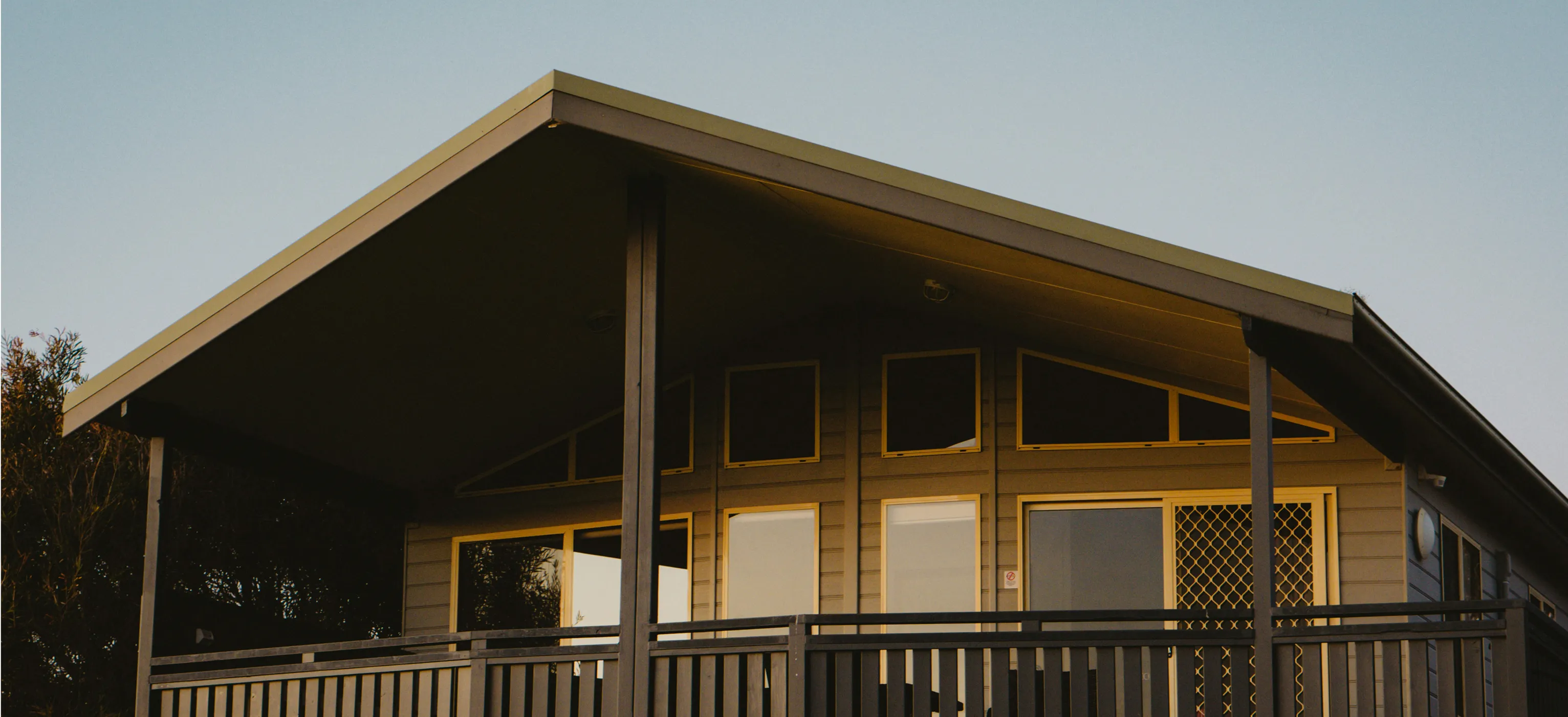 Modern house with beige siding, large windows, and a covered porch with railing at sunset.