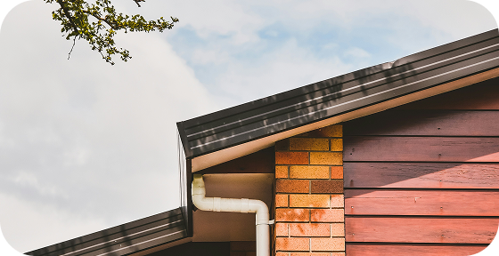 Close-up of a house roof corner with a white gutter downspout against a partly cloudy sky.