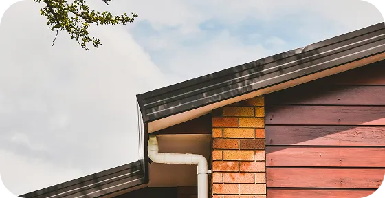 Close-up of a house roof corner with a white gutter downspout against a partly cloudy sky.