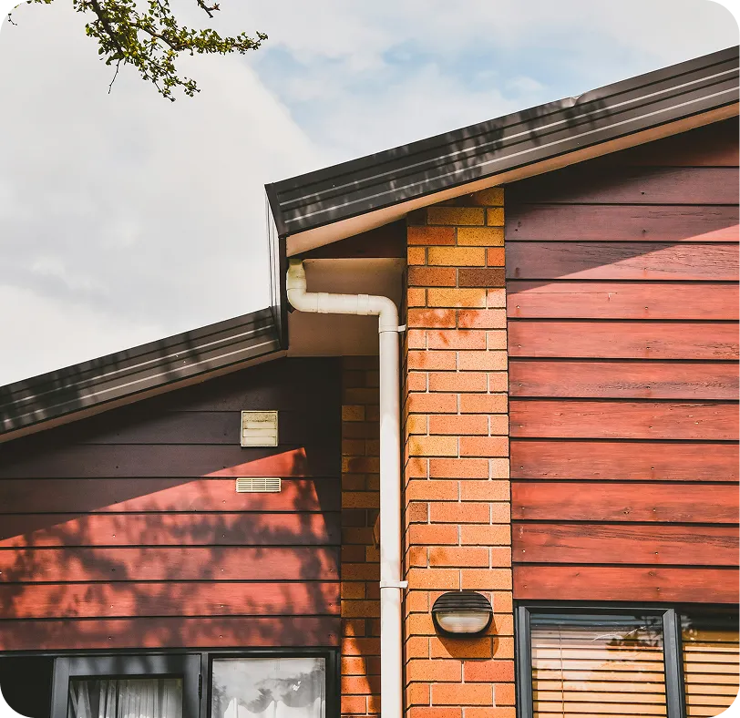 Section of a house exterior showing a slanted roof, brick and wooden siding, white gutter pipe, and windows with blinds and curtains.