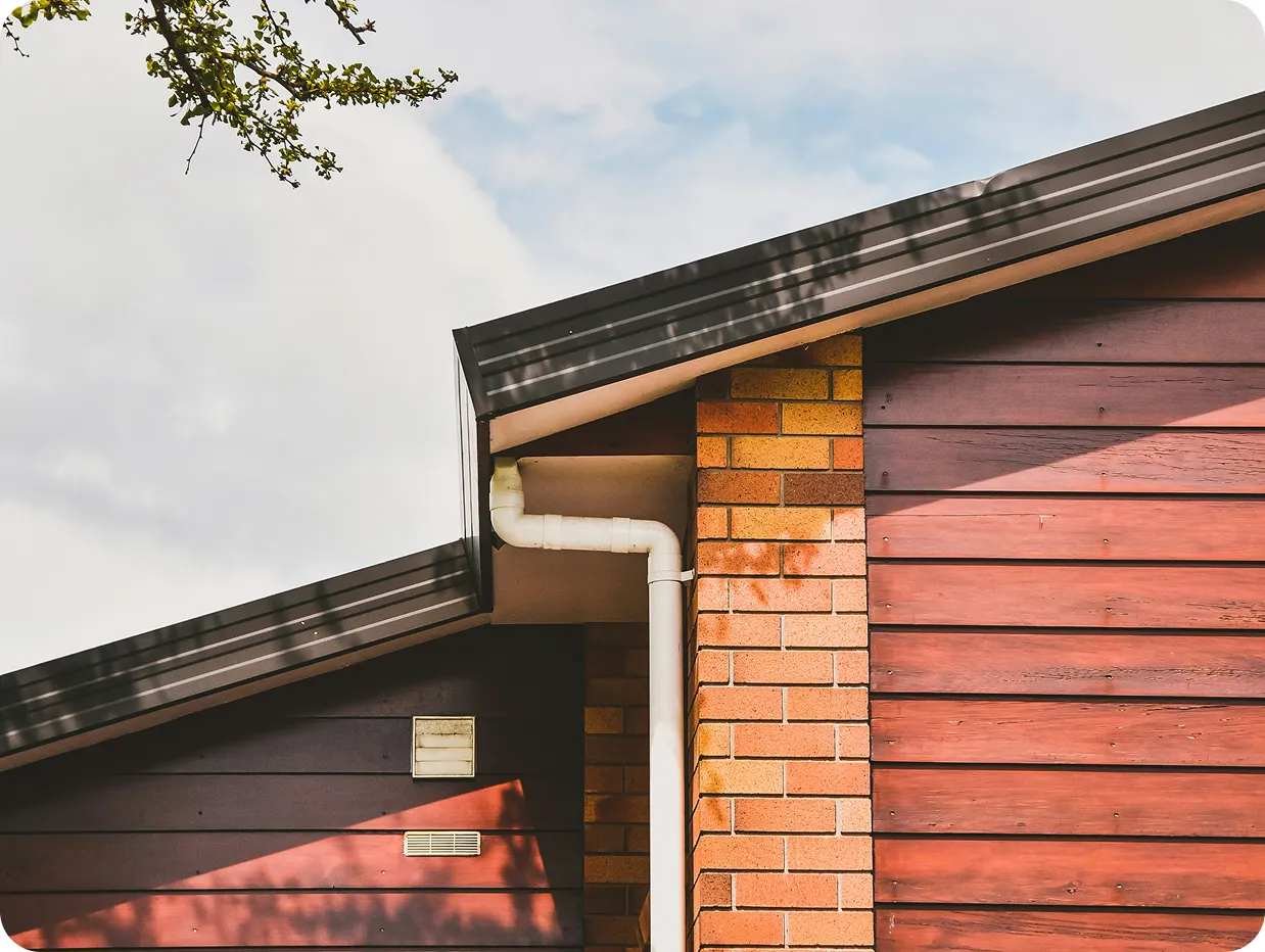 Side view of a house corner with red wooden siding, yellow brick column, white rain gutter, and cloudy sky background.