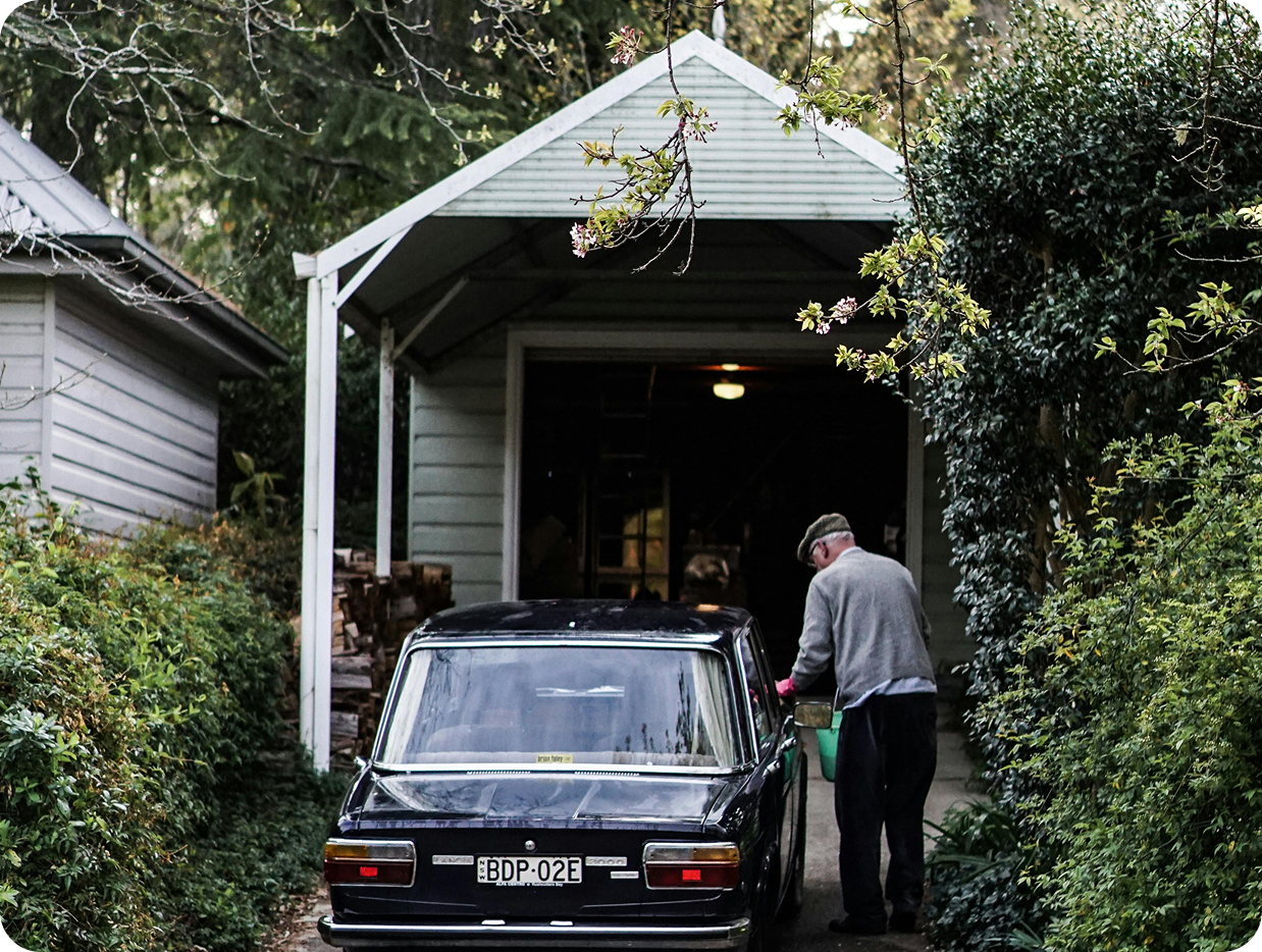 Older man wearing a flat cap cleaning or inspecting the side of a black vintage car parked in front of a small white garage surrounded by greenery.