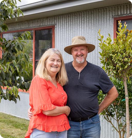 Smiling middle-aged couple standing outside near trees and a house with corrugated metal siding.