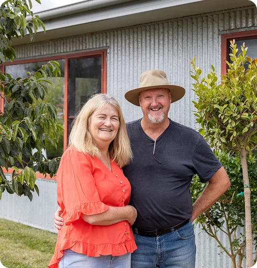 Smiling middle-aged couple standing outside near trees and a house with corrugated metal siding.