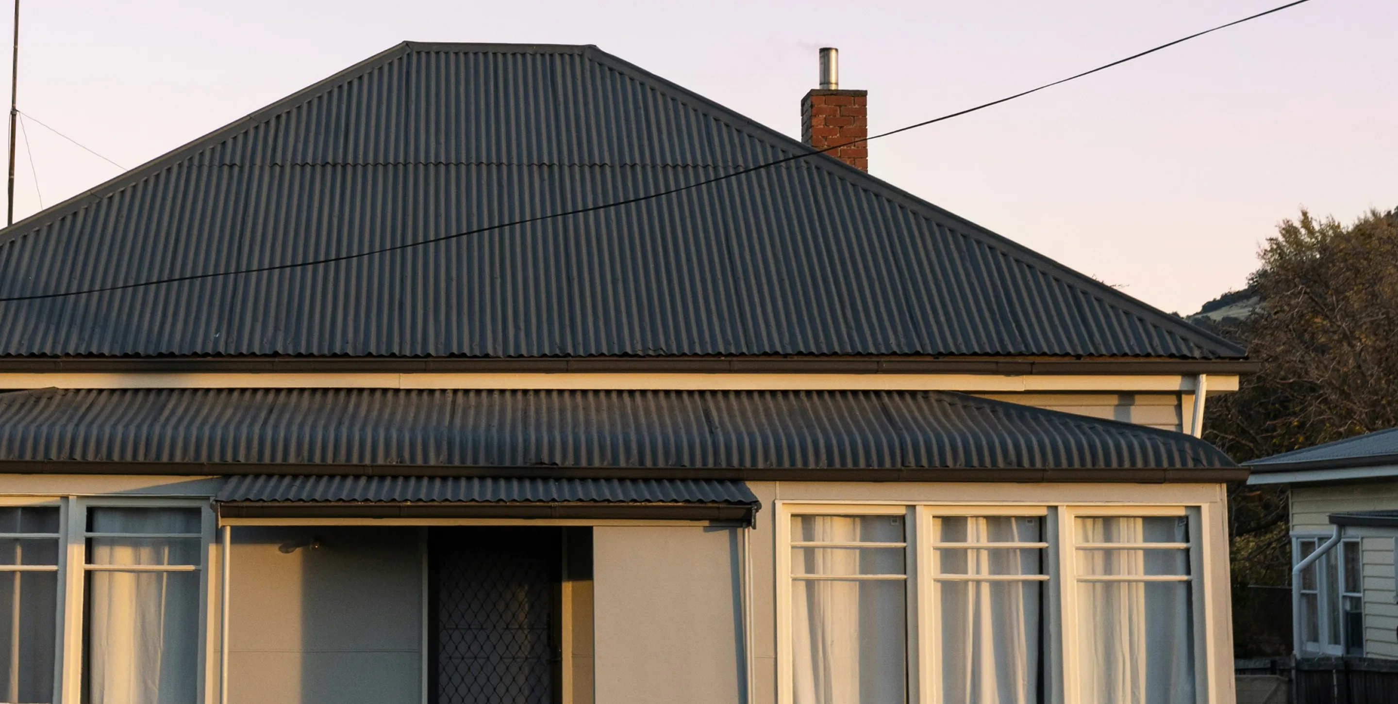 House with corrugated metal roof, chimney, and white-framed windows with curtains during sunset.