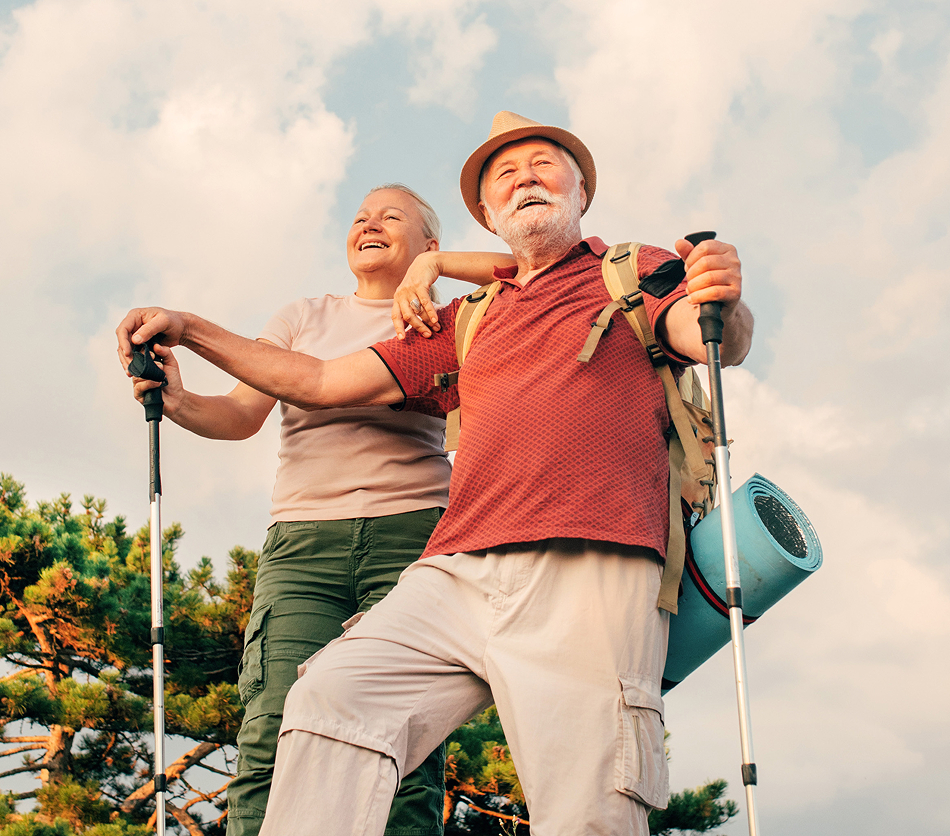 Smiling elderly couple hiking outdoors with walking poles under a partly cloudy sky.