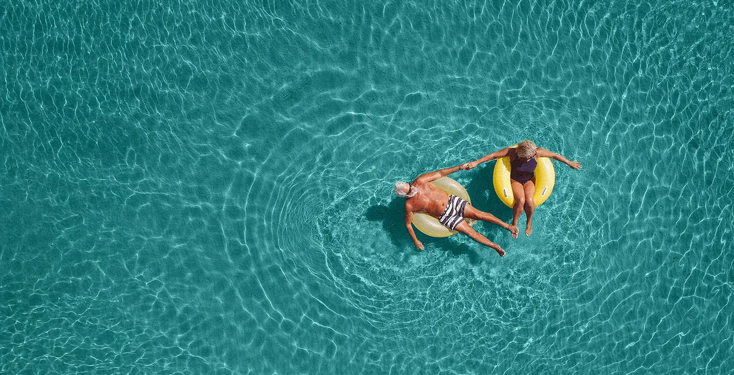 A man and a woman holding hands while floating on yellow inflatable rings in clear blue water.