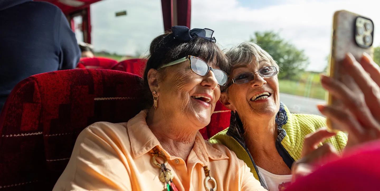 Two elderly women smiling and taking a selfie together on a bus with red seats and a window showing a green landscape outside.