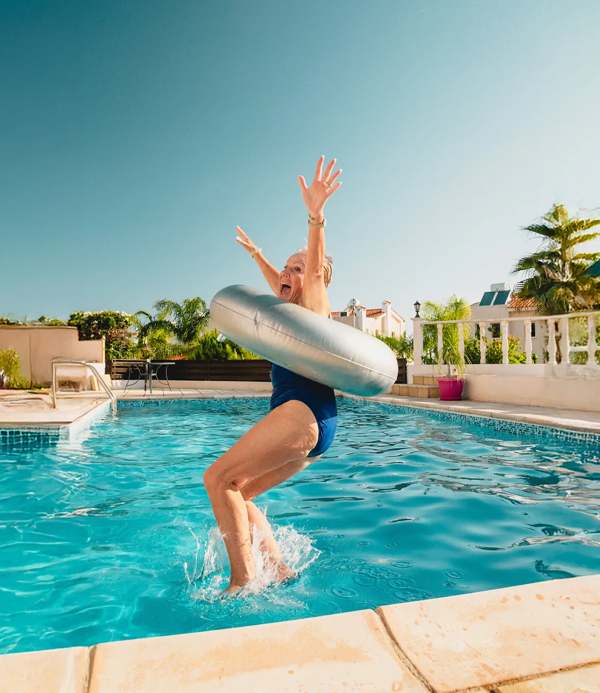 Elderly woman joyfully jumping into a pool wearing a blue swimsuit and a silver inflatable ring.