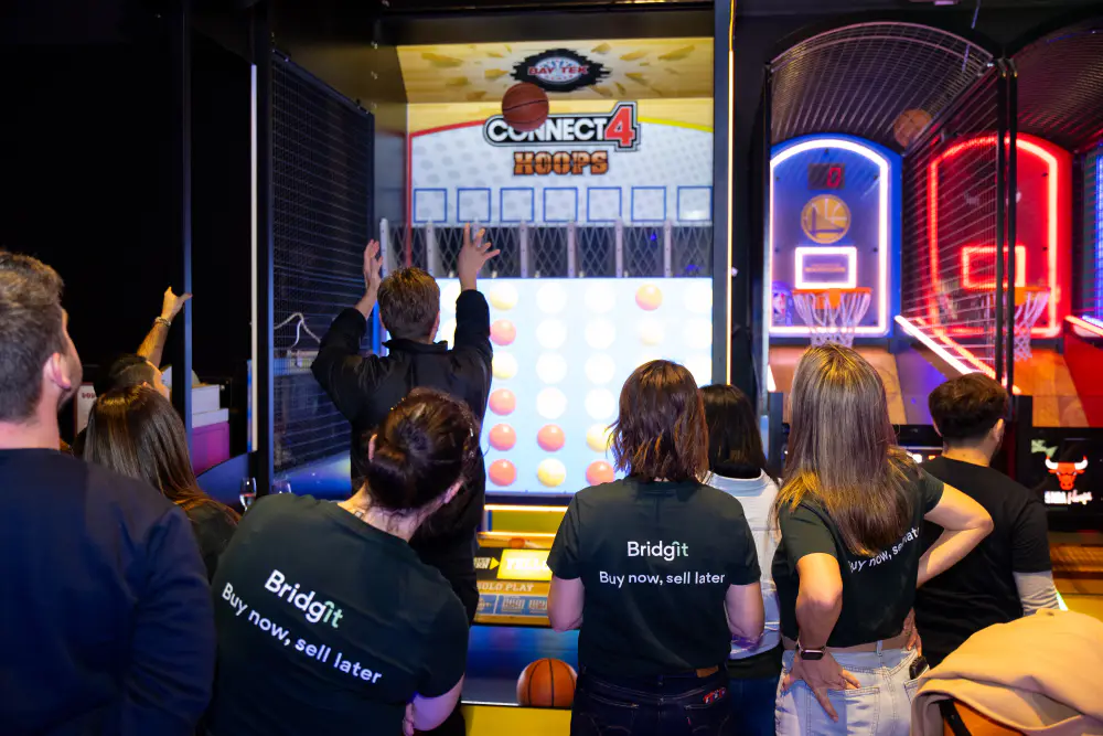 Bridgit team members in branded T-shirts cheering on a colleague at an arcade basketball game