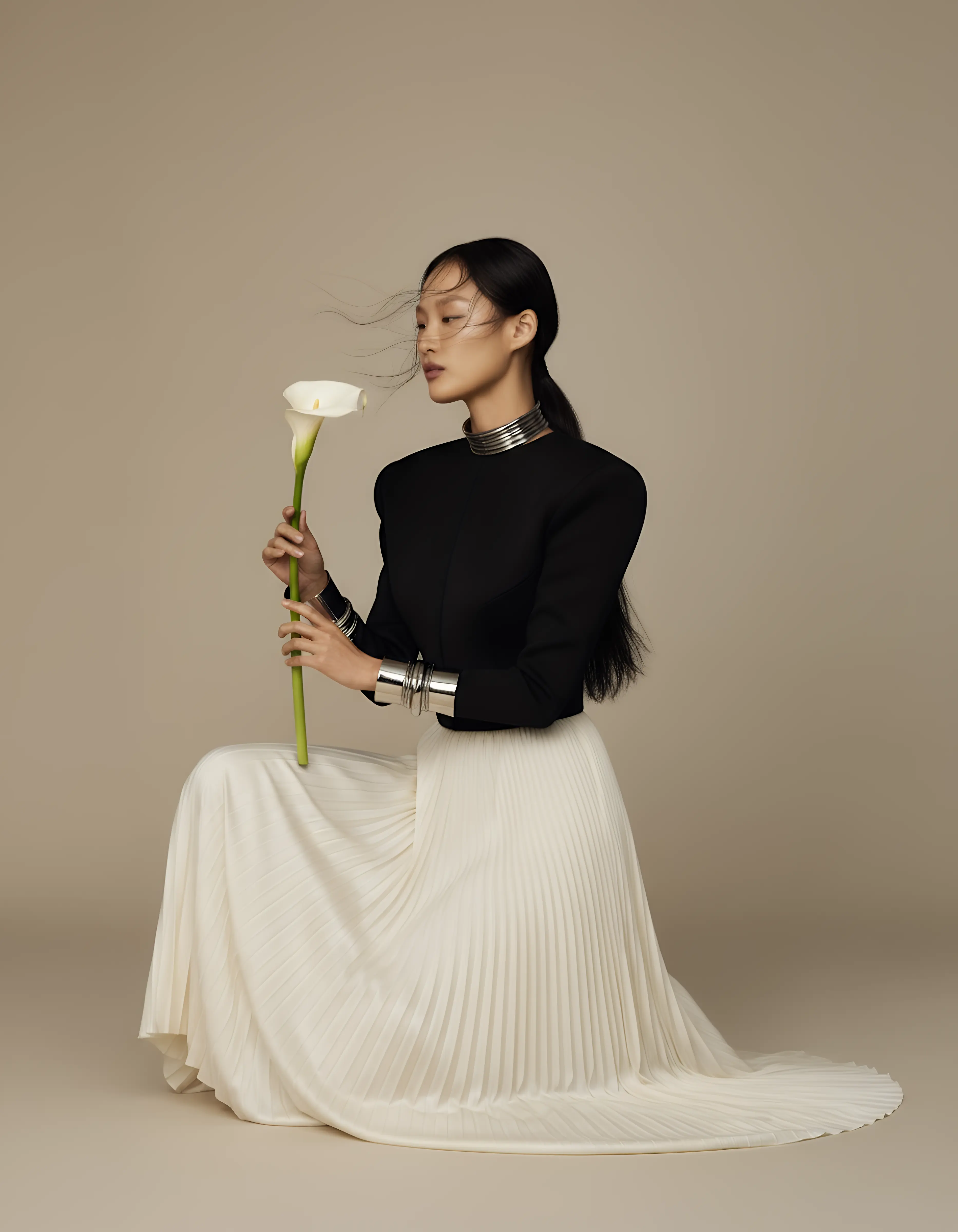 Elegant woman in a black top and white pleated skirt kneeling and holding a white calla lily against a beige background.