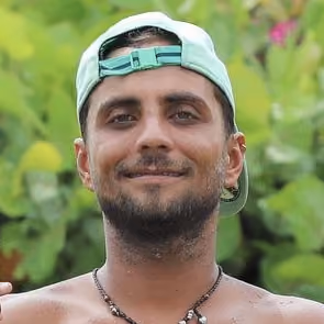 Homme souriant portant une casquette verte à l'envers, collier, avec un fond de feuillage vert flou.