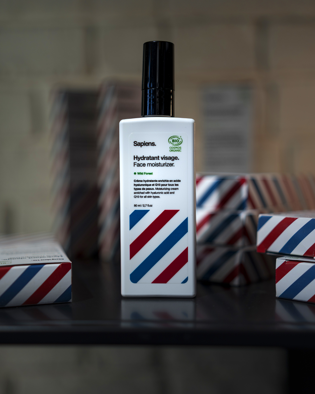 Bouteille blanche de crème hydratante pour le visage Sapiens avec dessin de bandes rouges et bleues, entourée de boîtes assorties en arrière-plan flou.