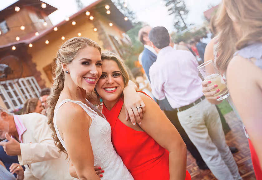 Bride in lace dress hugging a friend in red during an outdoor party at Hacienda La Danesa.