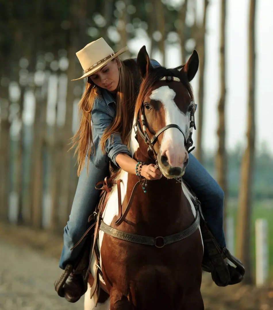 A woman wearing a hat and a denim shirt rides a brown and white pinto horse on a tree-shaded path. An exclusive equestrian experience at Hacienda La Danesa.
