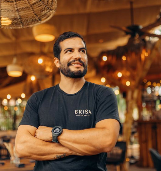 A friendly and welcoming staff member at Hacienda La Danesa, wearing a black Brisa logo t-shirt, posing with arms crossed in the warm, sunlit atmosphere of the hotel's restaurant.