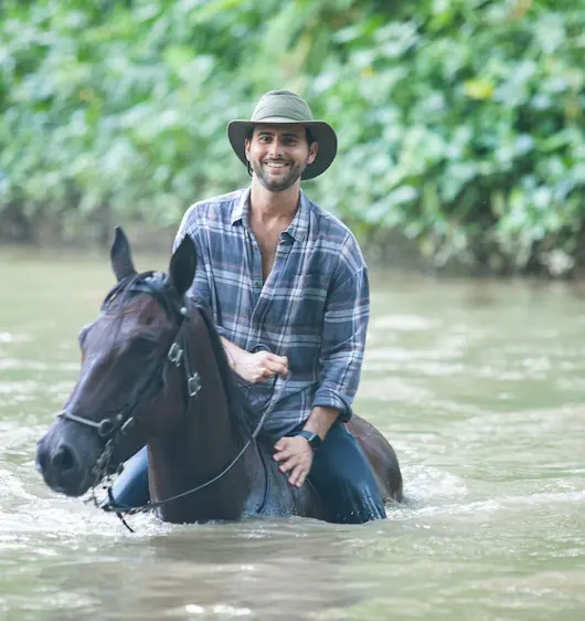 Un hombre sonriente con sombrero de ala ancha y camisa de cuadros monta un caballo oscuro a través de aguas poco profundas, rodeado de exuberante vegetación verde.