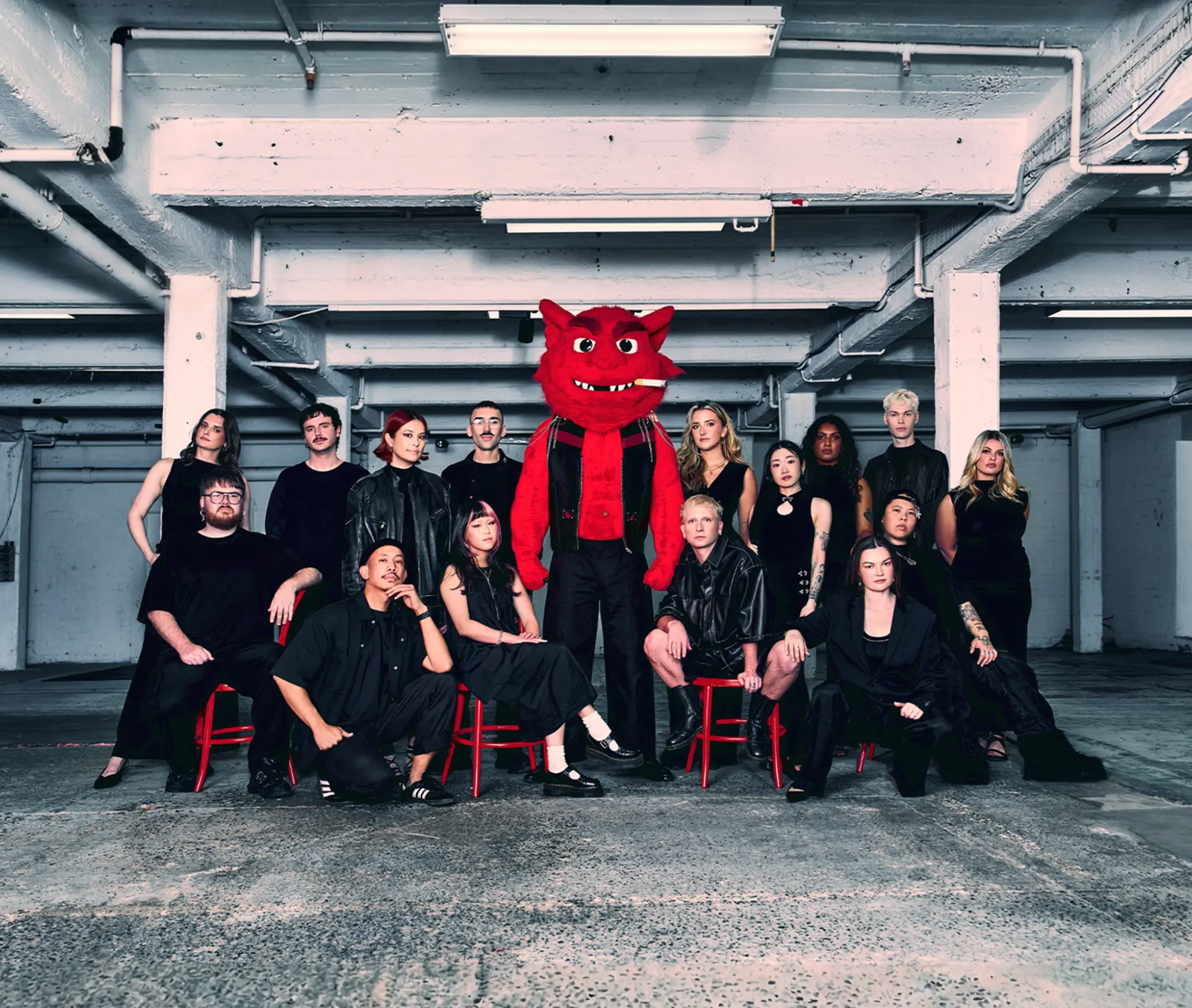 Group portrait of diverse young adults wearing black clothing with one person in the center dressed in a red devil costume smoking a cigarette, posed in an industrial space.