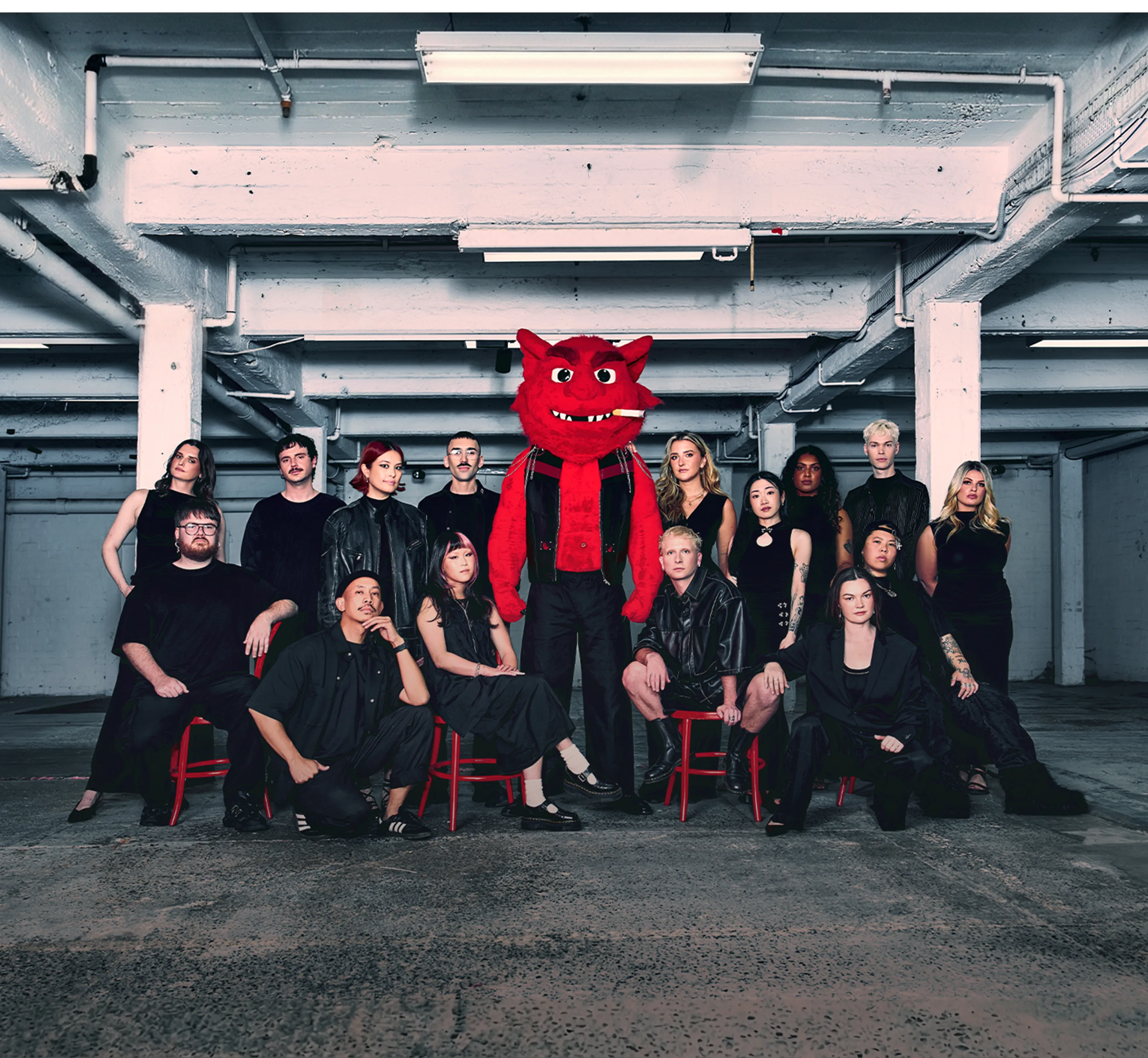 Group of diverse people dressed mostly in black, with one person in a red devil costume in an industrial space.