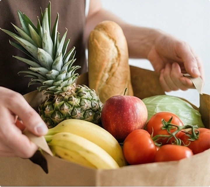 Person hält Papiertüte mit Ananas, Bananen, Apfel, Tomaten, Kopfkohl und Baguettebrot.