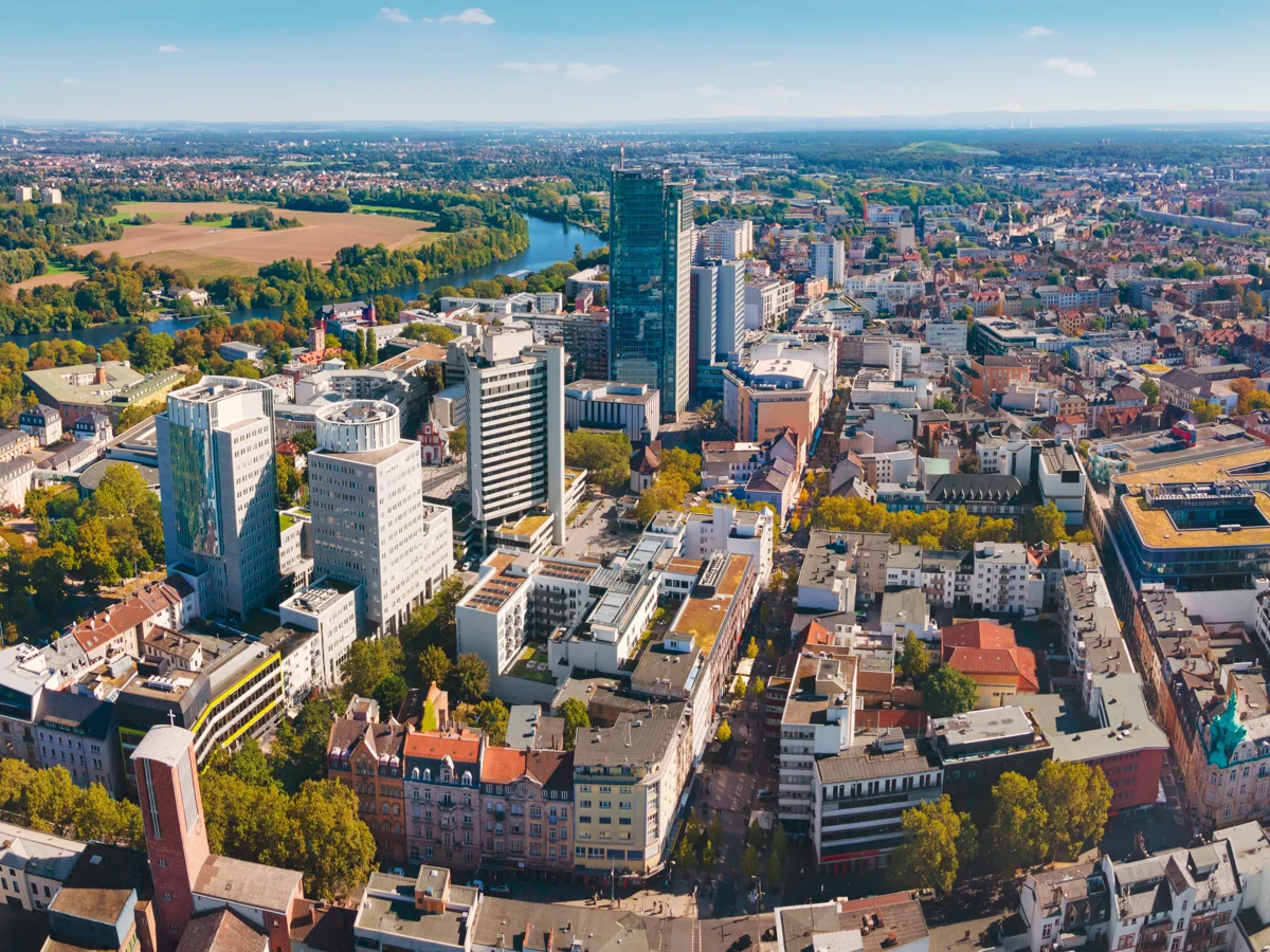 Luftaufnahme einer Stadt mit Hochhäusern, Fluss und weitläufigem grünen Umland bei klarem Himmel.
