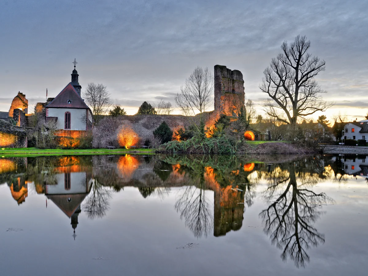 Ruinen und Bäume spiegeln sich bei Sonnenuntergang in einem ruhigen See.