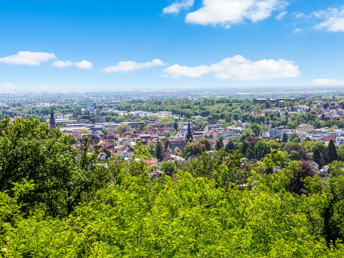 Blick auf eine Stadt mit Kirchtürmen und vielen Bäumen unter blauem Himmel mit weißen Wolken.
