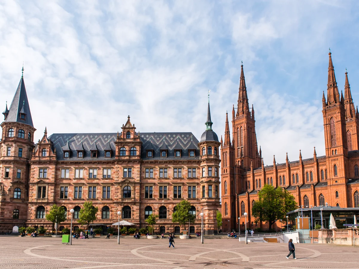 Historisches Gebäude und rote Kirchenfassade mit Türmen unter blauem Himmel auf einem Platz mit Bäumen