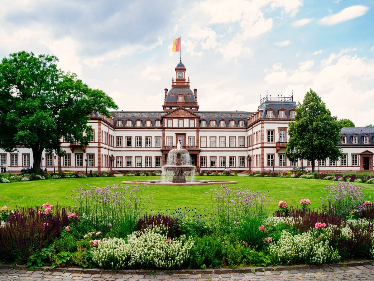 Schloss mit zentralem Brunnen, Blumenbeeten, großen Bäumen und einer Flagge auf dem Dach.