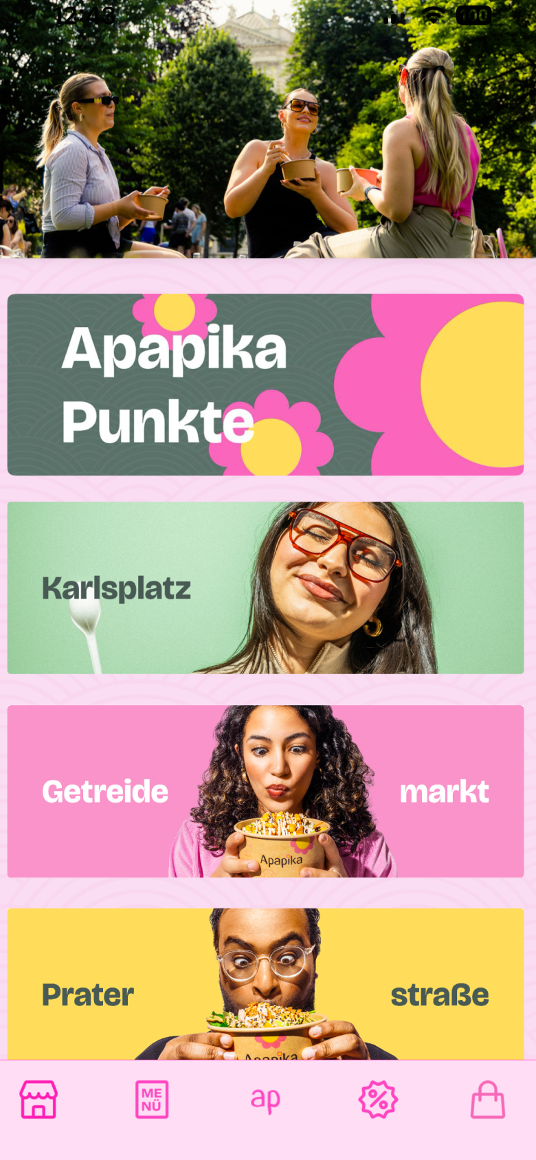 Drei Personen essen draußen in einem Park, darunter zwei Frauen mit Sonnenbrillen, die zusammen sitzen und lachen.