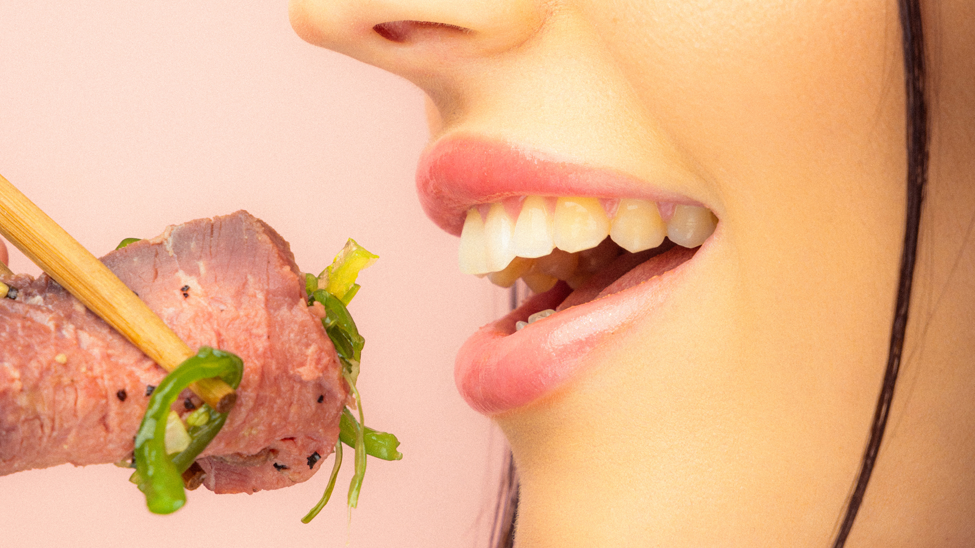 Nahaufnahme einer Person, die mit Stäbchen ein Stück Fleisch und grünes Gemüse zum Mund führt.