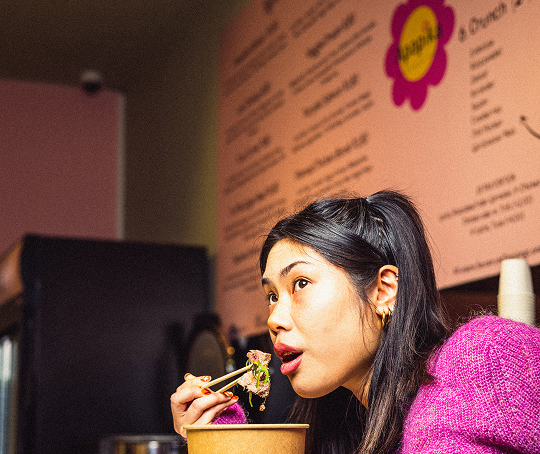 Frau in pinkem Pullover isst mit Stäbchen aus einem Pappbecher in einem Restaurant.