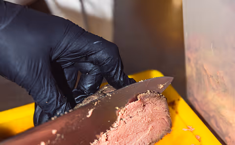 Hand mit schwarzem Handschuh schneidet gebratenes Fleisch auf einem gelben Schneidebrett mit einem großen Messer.