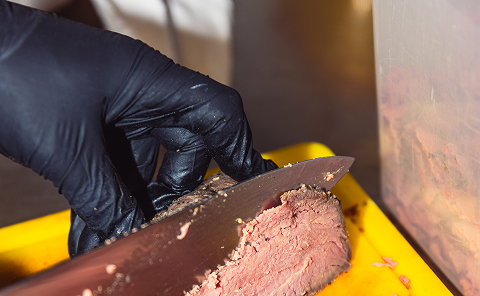 Hand mit schwarzem Handschuh schneidet gebratenes Fleisch auf einem gelben Schneidebrett mit einem großen Messer.