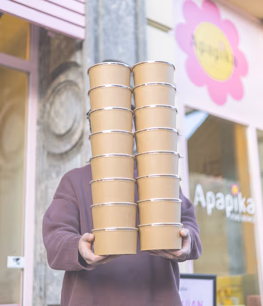 Person trägt hohen Stapel leerer brauner Pappbecher vor einem Geschäft mit Apapika-Schild.
