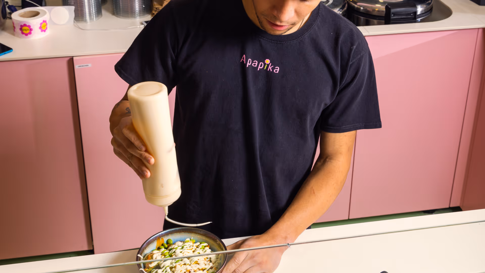 Mann in schwarzem Apapika-T-Shirt gibt cremige Sauce über eine Schüssel mit Gemüse in einer Küche mit rosa Schränken.