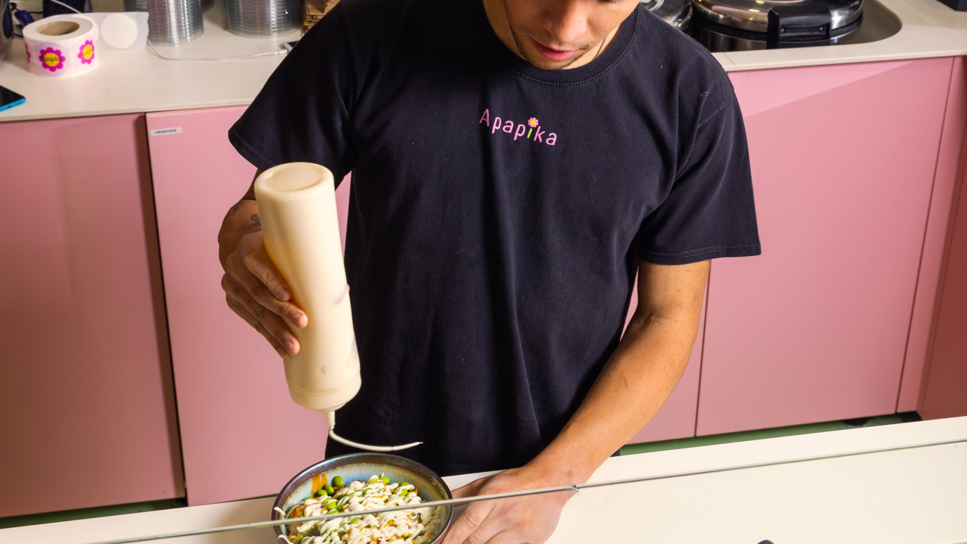 Mann in schwarzem Apapika-T-Shirt gibt cremige Sauce über eine Schüssel mit Gemüse in einer Küche mit rosa Schränken.