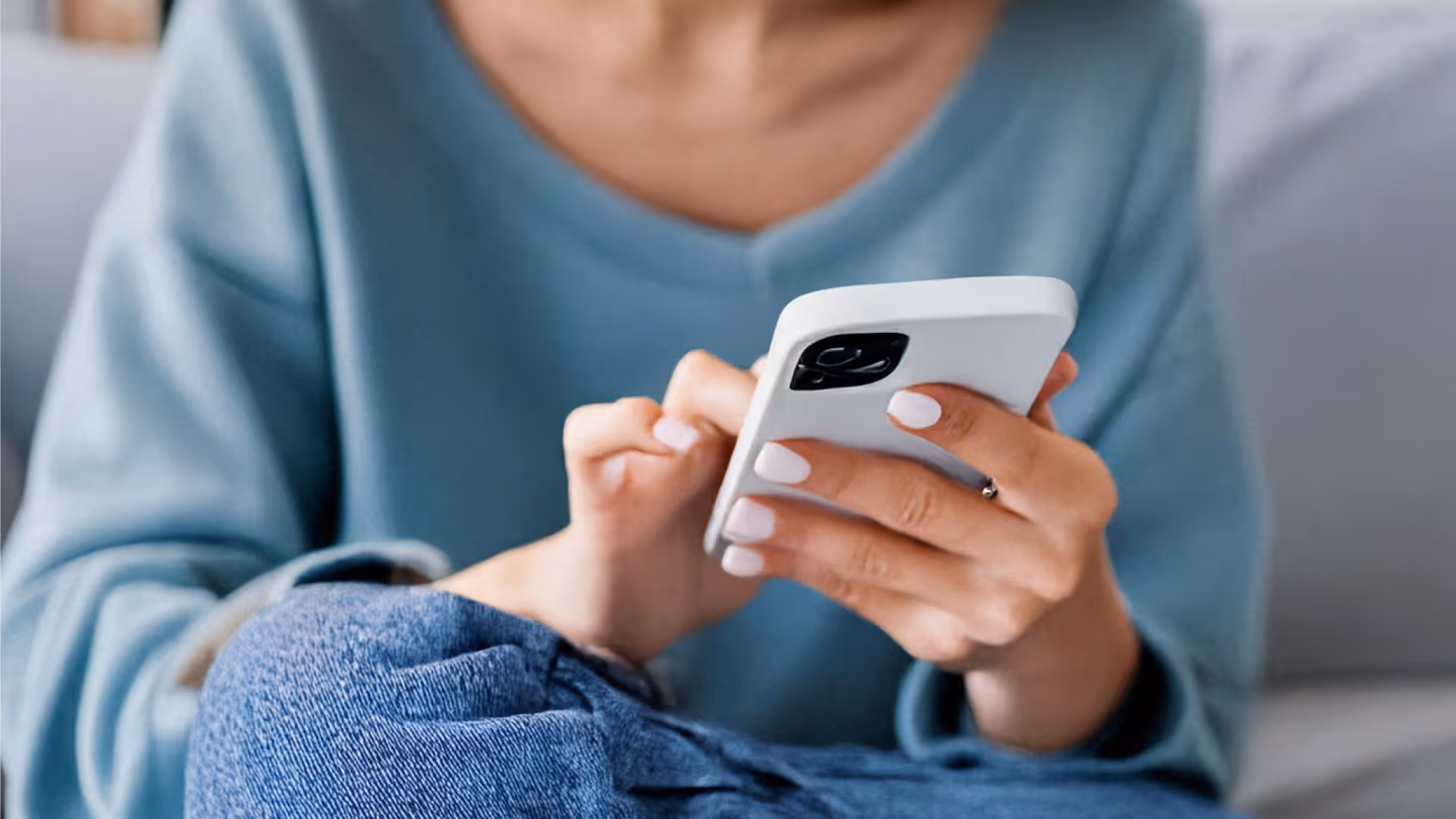 Person mit hellblauem Pullover, die ein weißes Smartphone hält und benutzt, auf einem Sofa sitzend.