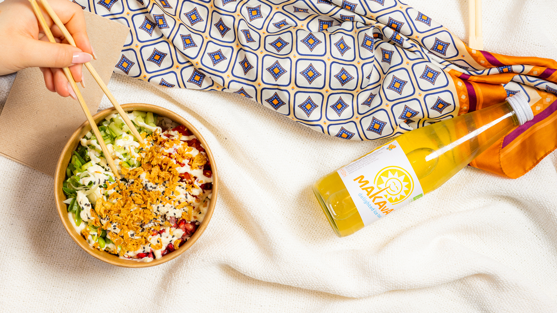 Person hält Essstäbchen über eine Schüssel mit frischem Gemüse, Sesam und knusprigen Toppings, daneben eine Flasche gelber Makava Eistee auf weißer Stoffdecke.