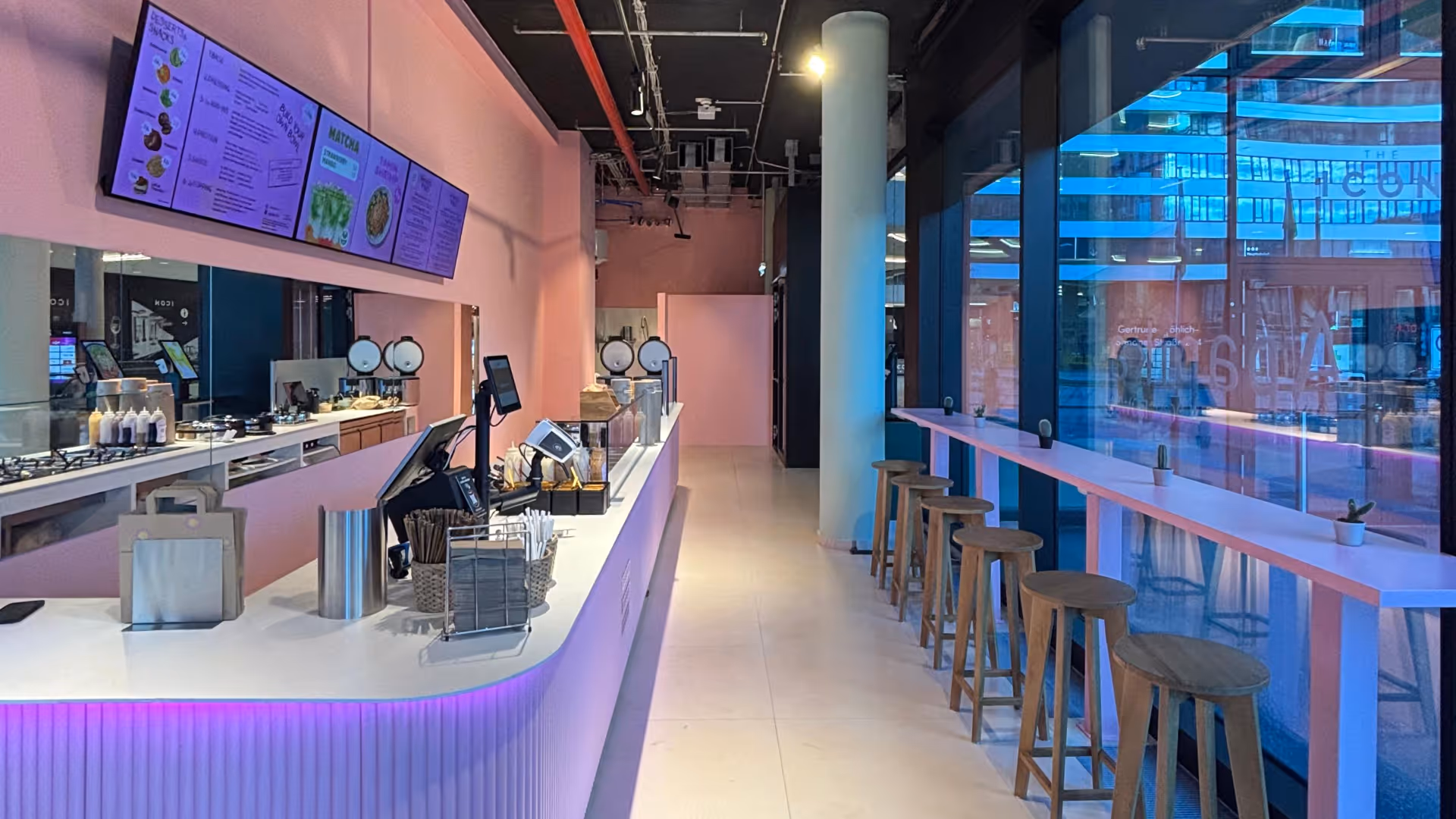 Modernes Café mit rosa Theke, Holzbarhockern und großen Fenstern zur Straßenseite.