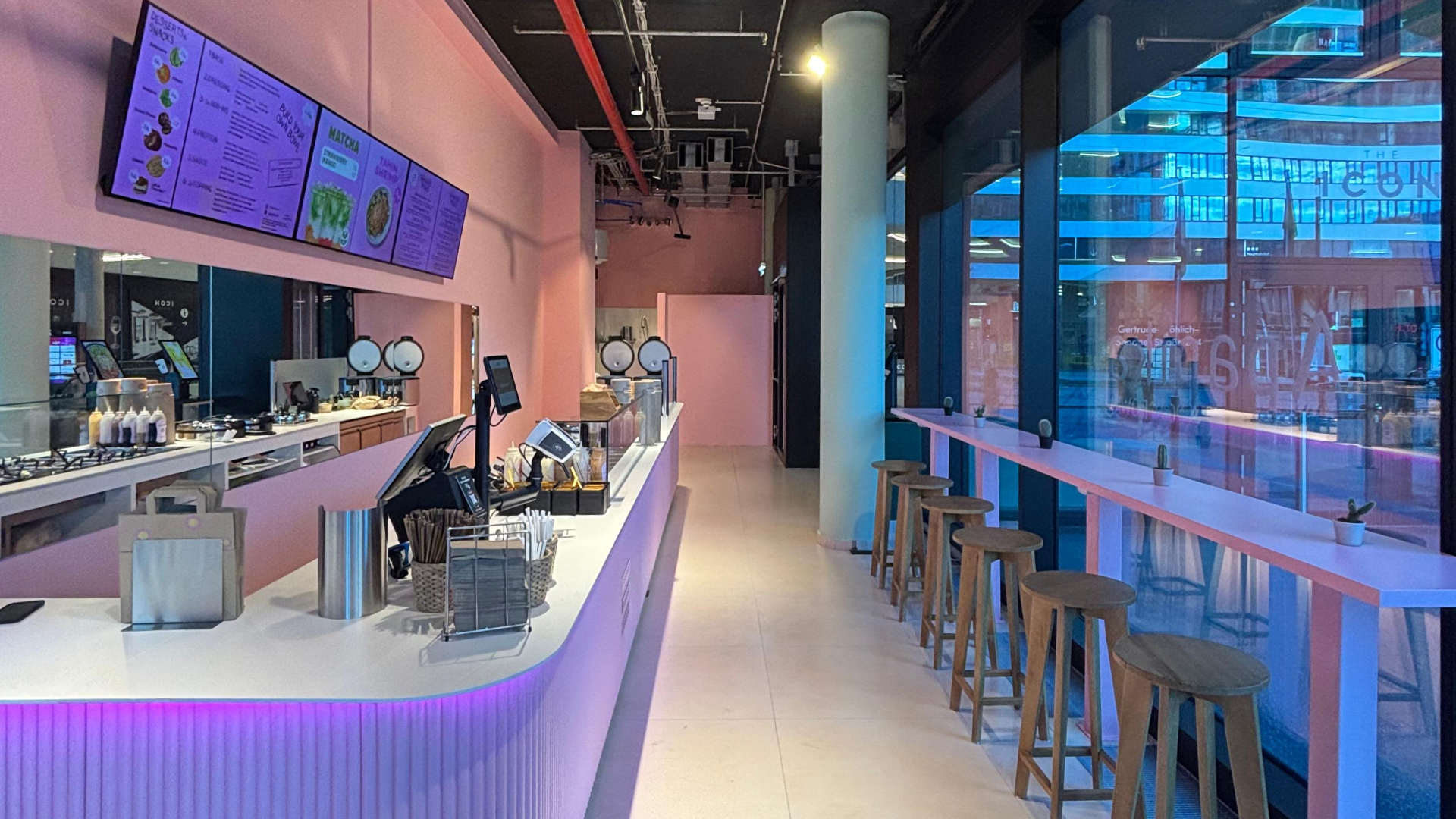 Modernes Café mit rosa Theke, Holzbarhockern und großen Fenstern zur Straßenseite.