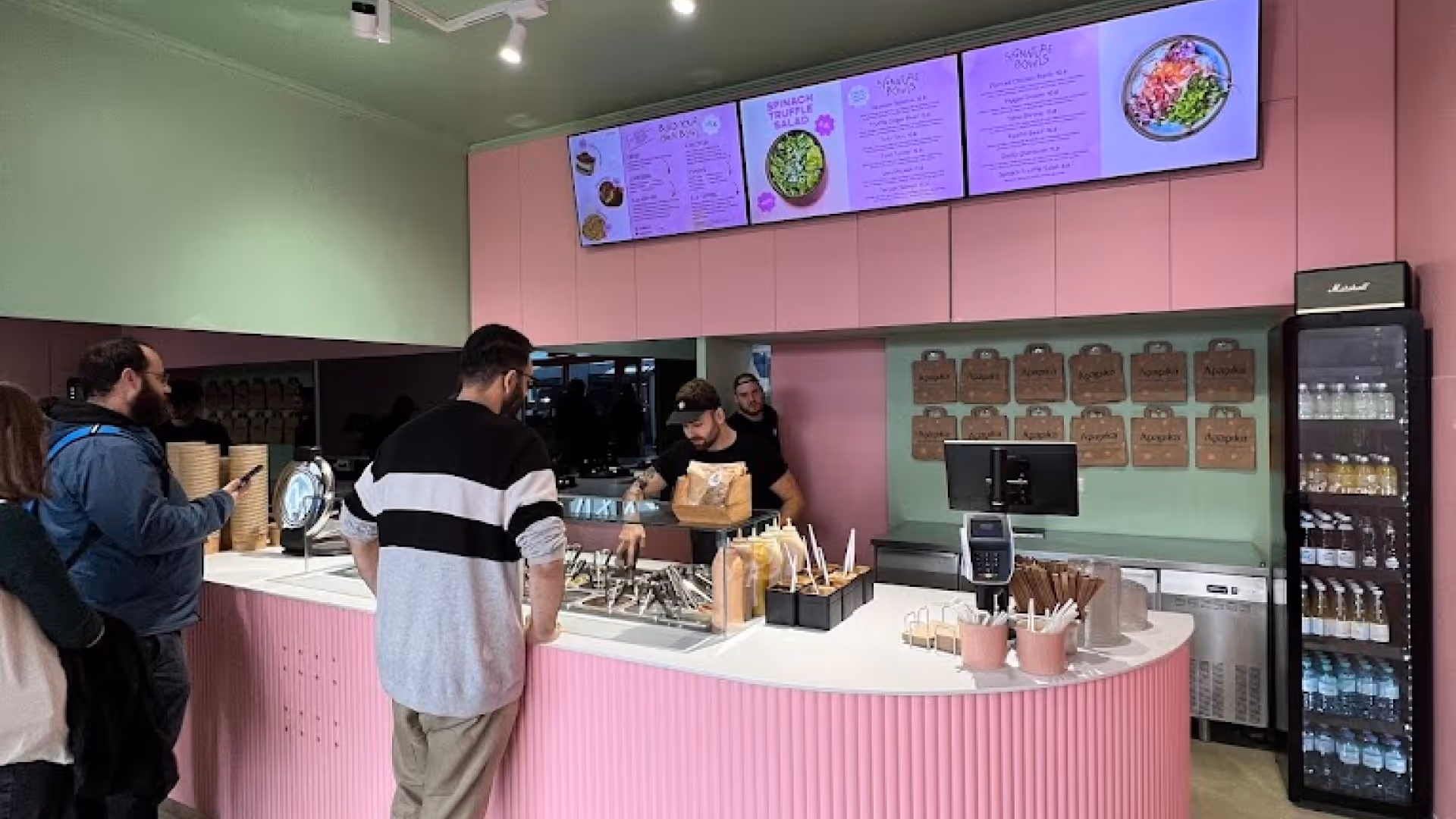 Innenansicht eines Restaurants mit rosa Theke, Kunden in der Warteschlange und digitalem Speisebildschirm über dem Tresen.