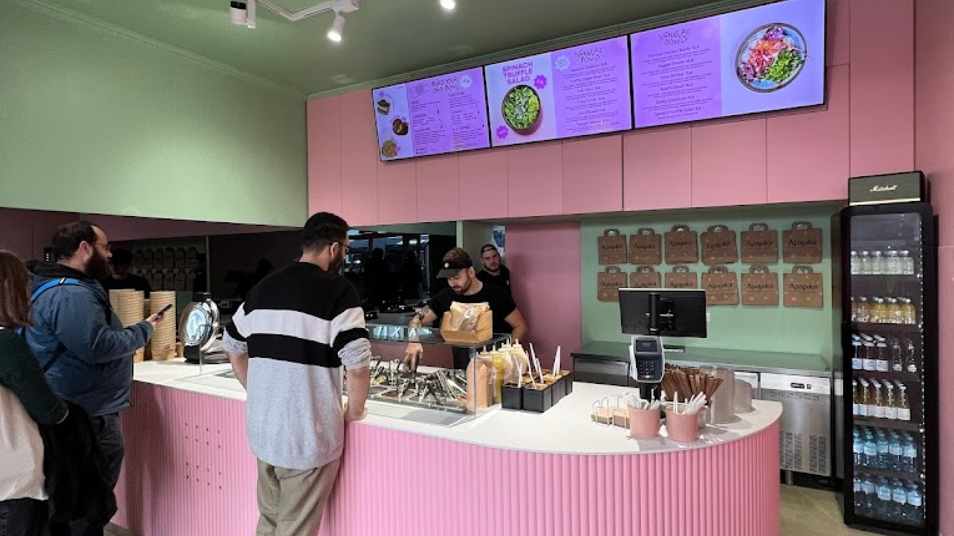 Innenansicht eines Restaurants mit rosa Theke, Kunden in der Warteschlange und digitalem Speisebildschirm über dem Tresen.