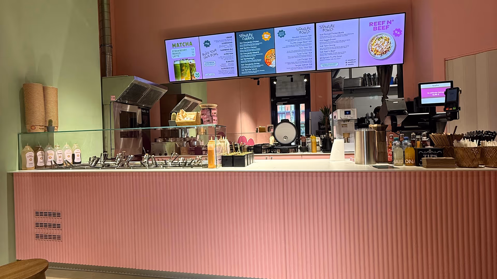 Theke eines modernen Gastronomiebetriebs mit rosa Front, verschiedenen Saucen, Strohhalmen, und digitalen Menüanzeigen an der Wand.