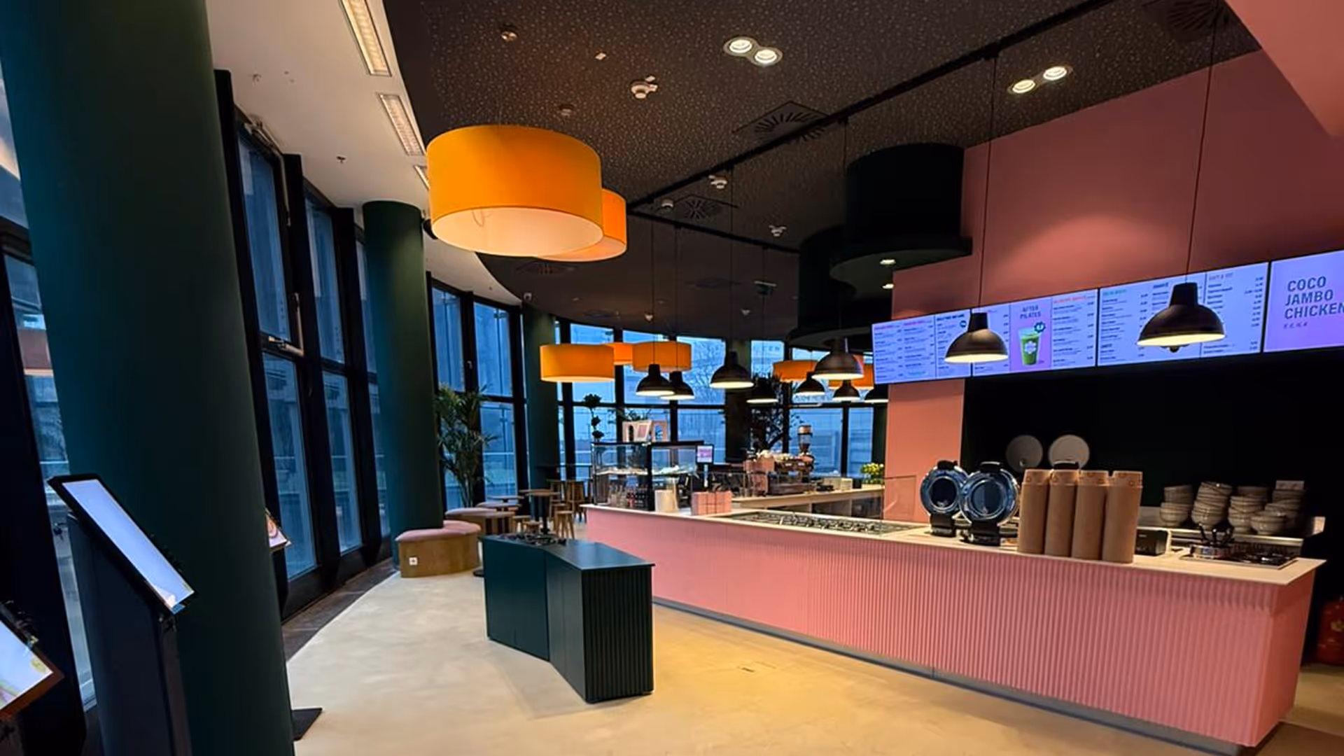 Modernes Café mit rosa Theke, orangefarbigen und schwarzen Hängeleuchten und großen Fenstern im Hintergrund.