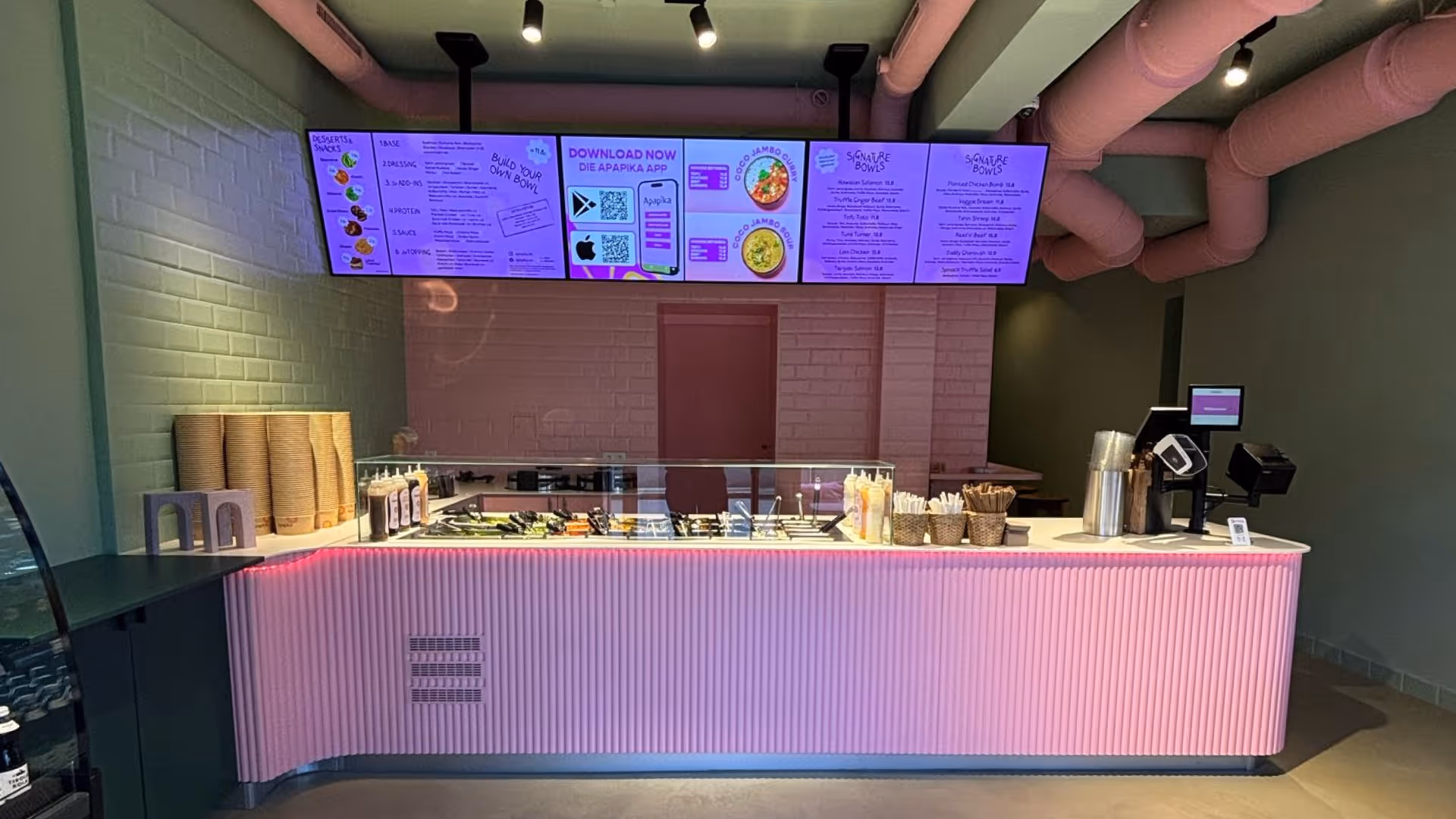 Restaurant mit modernem pink beleuchtetem Tresen, Salatbar und digitalen Menüs auf Bildschirmen unter rosa Deckenrohren.